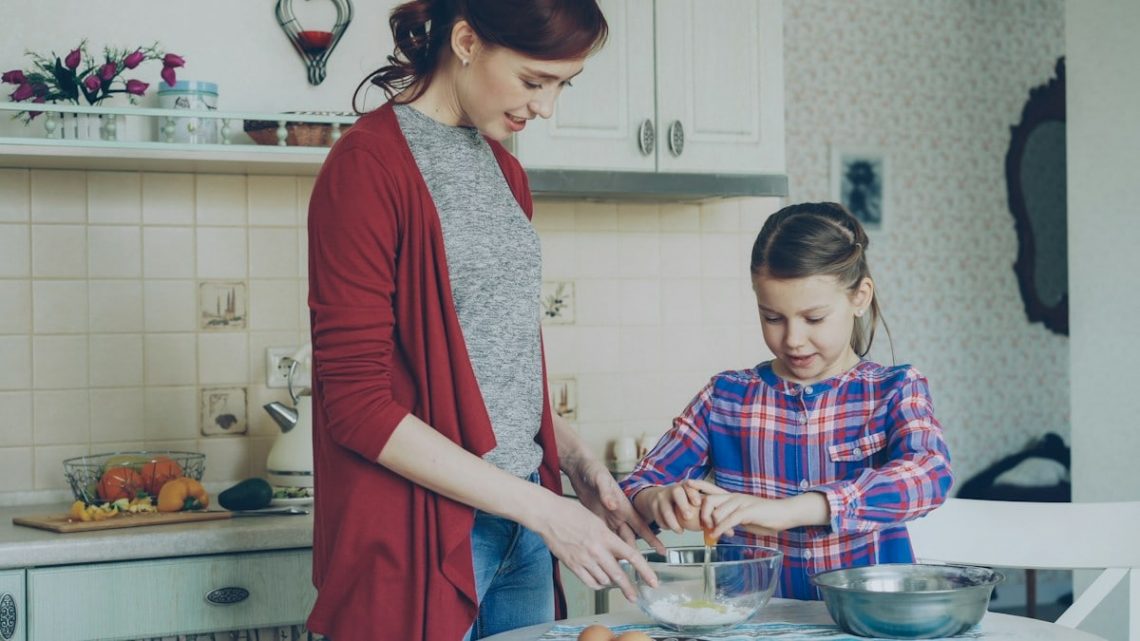 Little cheerful girl helping her mother in the kitchen cropping egg into bowl and mixing dough for cookies. Family, food, home and people concept