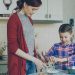 Little cheerful girl helping her mother in the kitchen cropping egg into bowl and mixing dough for cookies. Family, food, home and people concept